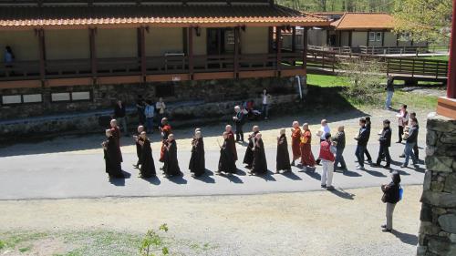 A line of monks in robes walking along a path near a building, with people seated and standing nearby.