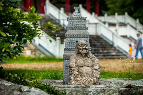 A stone statue of a seated Buddha beside a pagoda, surrounded by greenery and a blurred background of people walking.
