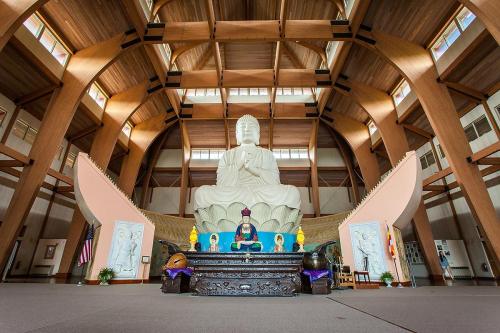 A spacious temple interior featuring a large seated Buddha statue, wooden beams, and decorative elements.