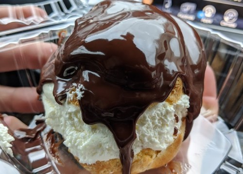 A close-up of a dessert with whipped cream and a glossy chocolate drizzle on top, held in a clear container.