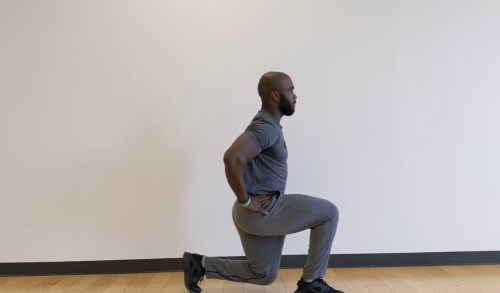 A person in athletic wear performs a lunge exercise, with one knee bent and hands on hips, against a plain wall.