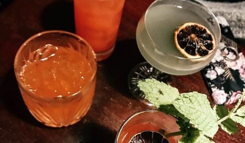 A top-down view of three colorful cocktails on a wooden table, garnished with mint and dried fruit.