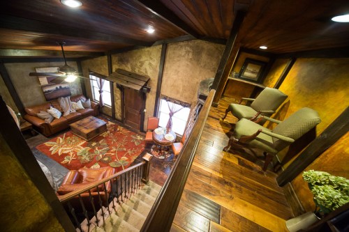 Cozy living room with wooden beams, leather sofas, a floral rug, and two armchairs, viewed from an upper level.