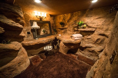 Cozy bathroom with rock-like walls, a mirror, a lamp, and a chair, featuring plants and soft brown carpeting.