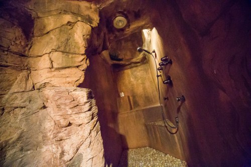 A rustic shower area with rock walls, featuring a showerhead and stone flooring, illuminated by soft lighting.