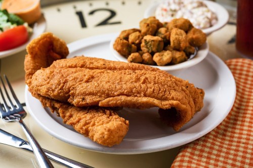 A plate of crispy fried fish fillets with a side of fried okra, served on a table with utensils and a checkered napkin.