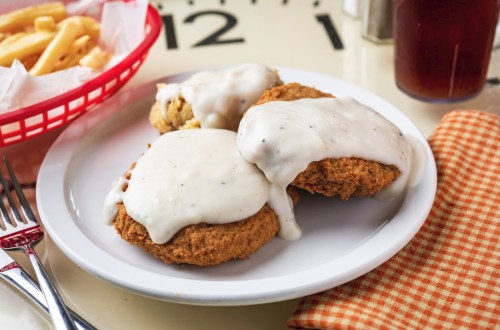 A plate of three fried chicken biscuits smothered in creamy white gravy, served with a side of fries and iced tea.