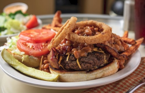 A hearty burger topped with bacon, cheese, and beans, served with onion rings, fries, lettuce, tomato, and pickles.