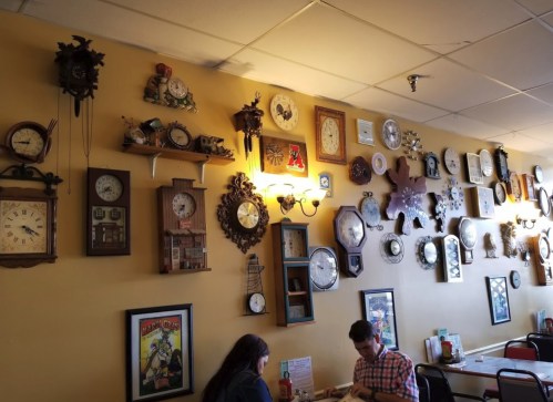 A cozy café interior featuring a wall adorned with various vintage clocks and two patrons seated at a table.