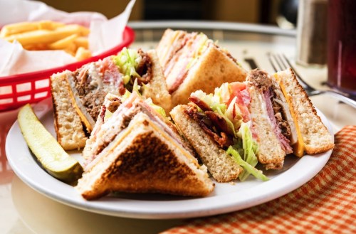 A plate of stacked sandwiches with layers of meat, cheese, and vegetables, served with a pickle and fries in the background.