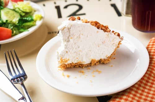 A slice of creamy pie with a graham cracker crust, topped with chopped nuts, on a white plate beside a salad.
