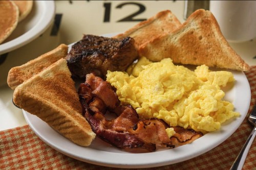 A plate of breakfast featuring scrambled eggs, bacon, toast, and a piece of steak on a checkered napkin.