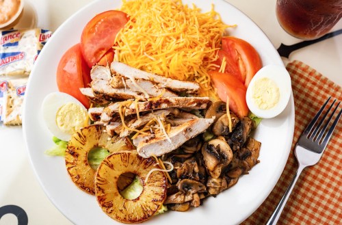 A plate of grilled chicken, sliced tomatoes, shredded cheese, mushrooms, pineapple, and hard-boiled eggs on a bed of lettuce.