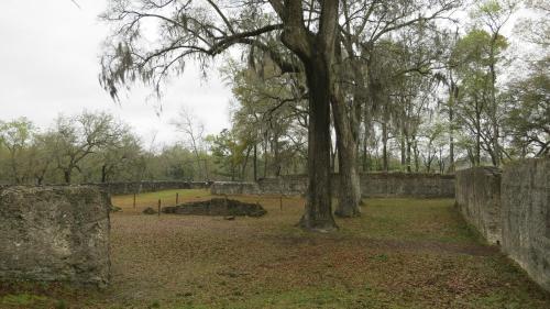 Ruins of an old structure surrounded by trees and Spanish moss, with a grassy area in the foreground.