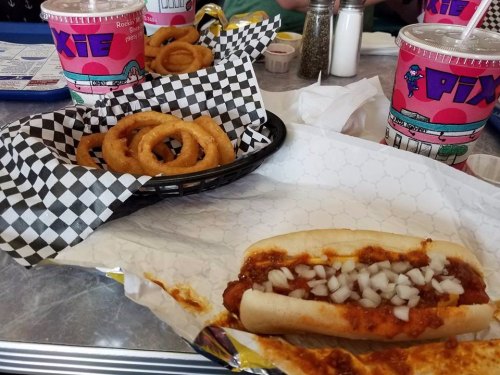A plate with a chili dog topped with onions, alongside a basket of onion rings and colorful drinks in the background.