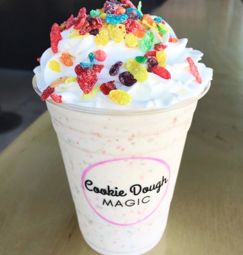 A creamy milkshake topped with whipped cream and colorful cereal sprinkles in a clear cup.