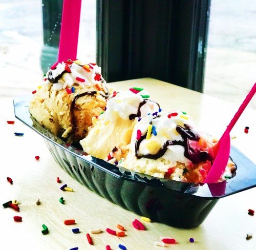 A colorful ice cream sundae in a black dish, topped with whipped cream, chocolate sauce, and sprinkles, with pink spoons.