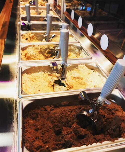 A variety of ice cream flavors displayed in metal tubs with scoops, set in a brightly lit ice cream shop.