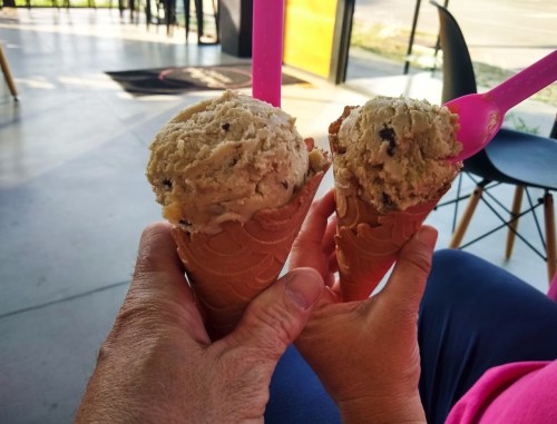 Two hands holding ice cream cones with cookie dough flavor, pink spoons visible, in a bright indoor setting.