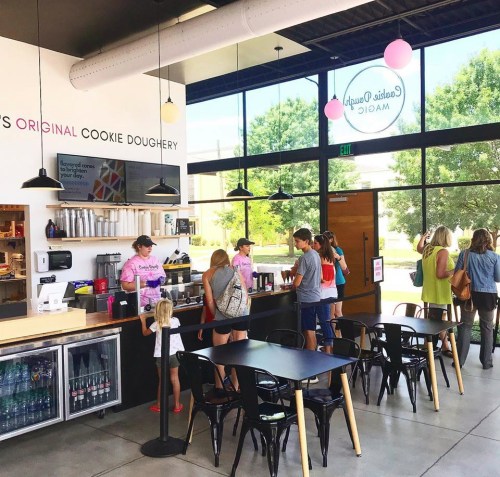 A busy cookie shop with customers at the counter, bright interior, and a menu displayed on the wall.