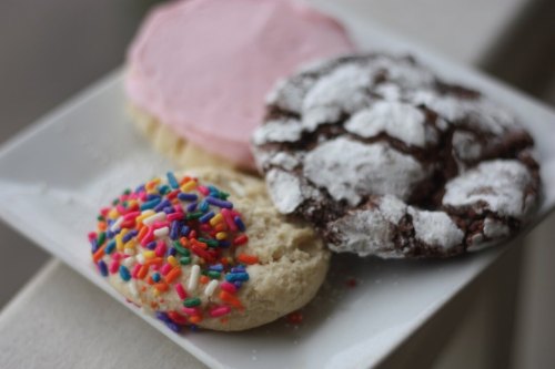 A plate with three cookies: one with pink frosting, one with colorful sprinkles, and a chocolate crinkle cookie.