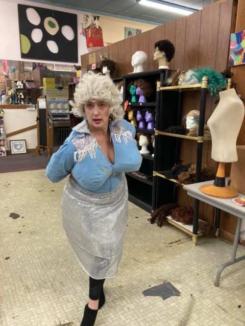 A person in a blue top and silver skirt poses in a quirky costume shop filled with wigs and mannequins.