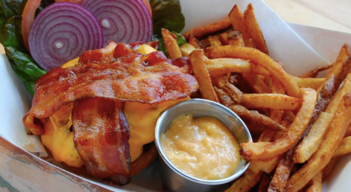 A juicy cheeseburger topped with crispy bacon, served with fries and a side of dipping sauce, garnished with onion slices.