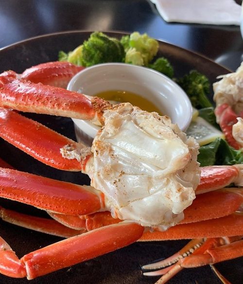 A close-up of a cooked crab with red claws, served on a plate with broccoli and a small bowl of dipping sauce.