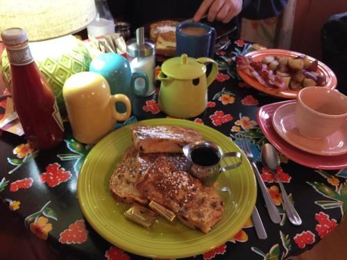 A colorful breakfast table featuring French toast, syrup, bacon, and various drinks, with a floral tablecloth.