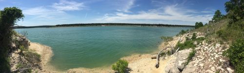 A panoramic view of a calm lake surrounded by rocky shores and lush greenery under a clear blue sky.