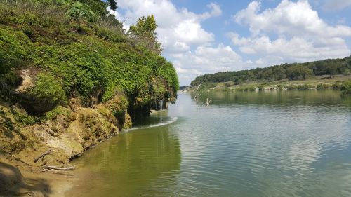 A serene river scene with lush green banks, clear water, and a blue sky dotted with fluffy clouds.