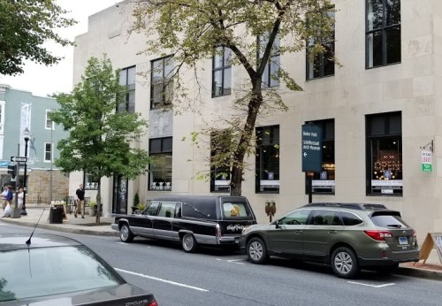 A street view featuring a black hearse parked beside a gray SUV in front of a modern building with large windows.