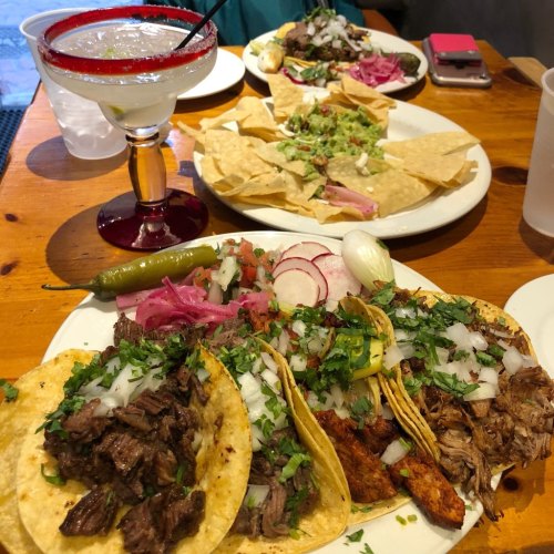 A table with tacos topped with onions and cilantro, a plate of nachos, and a margarita in a glass.