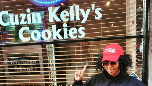 A person wearing a red cap and sunglasses poses in front of a shop window that reads "Cuzin Kelly's Cookies."