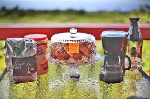 A glass cake stand with baked goods, coffee maker, coffee, and creamer on a table with a green landscape in the background.