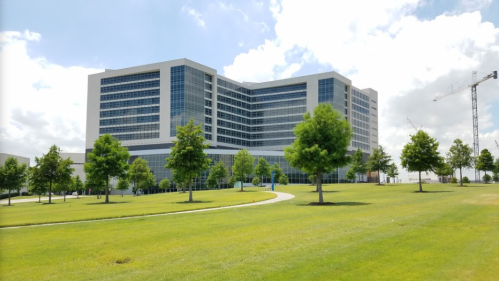 Modern multi-story building surrounded by green lawns and trees, with a construction crane in the background.