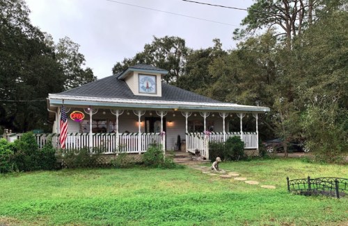 A charming house with a porch, an "Open" sign, and an American flag, surrounded by greenery and trees.