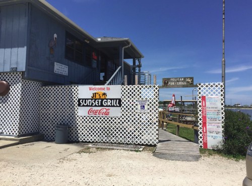 Entrance to JT's Sunset Grill, featuring a sign and a white lattice fence, with a clear blue sky above.