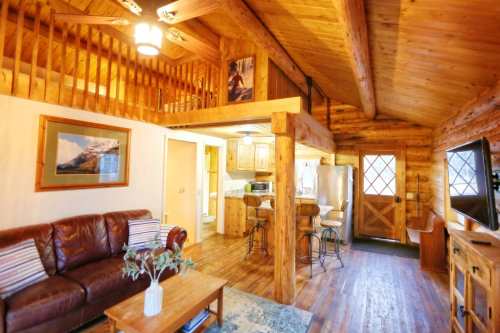 Cozy wooden cabin interior with a living area, kitchen, and loft, featuring rustic decor and warm lighting.