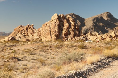 Rocky formations rise against a clear sky in a desert landscape, with sparse vegetation and distant mountains.