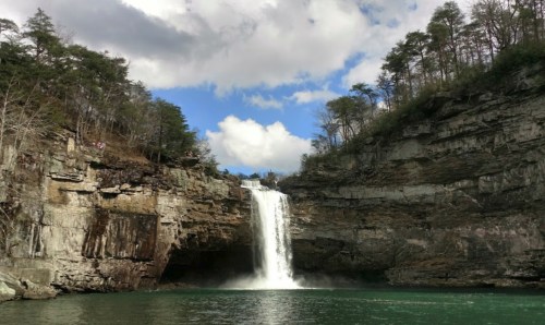 A stunning waterfall cascades into a serene pool, surrounded by rocky cliffs and lush trees under a partly cloudy sky.