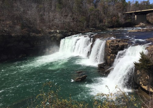 A scenic view of a waterfall cascading into a turquoise river, surrounded by trees and a bridge overhead.