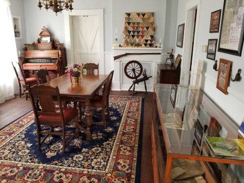 A cozy room with a wooden dining table, chairs, an antique organ, a spinning wheel, and a display case.