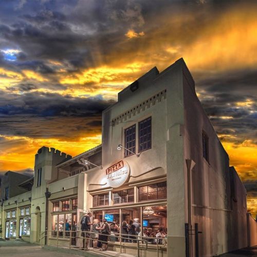 A pizza restaurant with people dining inside, set against a dramatic sunset sky with vibrant clouds.