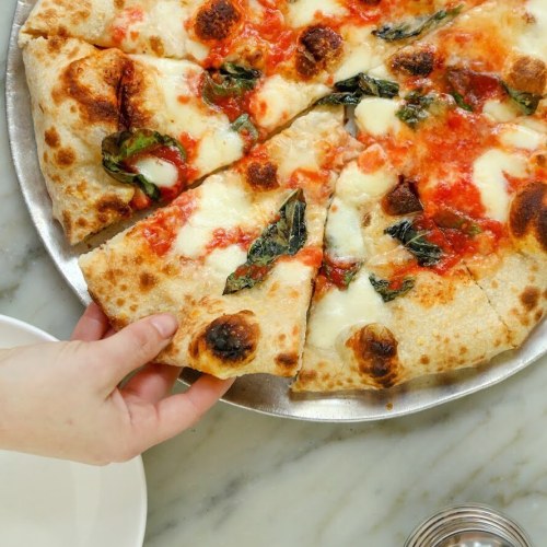 A hand reaching for a slice of pizza topped with mozzarella and basil on a silver platter.