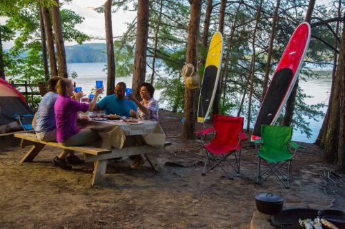 A group of friends enjoying a meal at a campsite by the lake, with kayaks and chairs nearby.