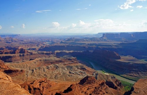A vast canyon landscape with layered rock formations and a winding river under a blue sky with scattered clouds.