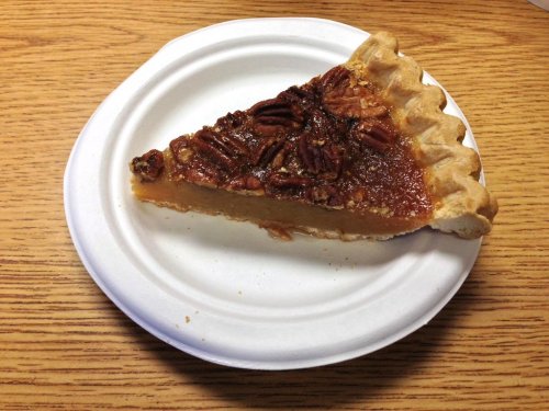 A slice of pecan pie on a white paper plate, featuring a golden filling and topped with whole pecans.