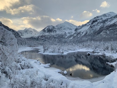 A serene winter landscape featuring snow-covered mountains, a calm river, and a cloudy sky.