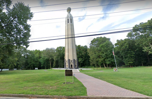 A tall monument surrounded by trees, with a paved path leading to it and a flagpole nearby.